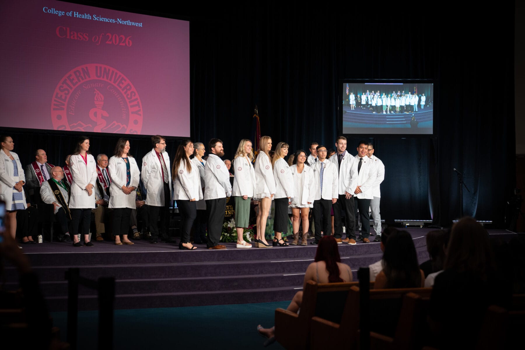 A group of people in white coats stands on a stage under the "Class of 2026" banner at a College of Health Sciences event, with an audience seated in the foreground and a display screen.