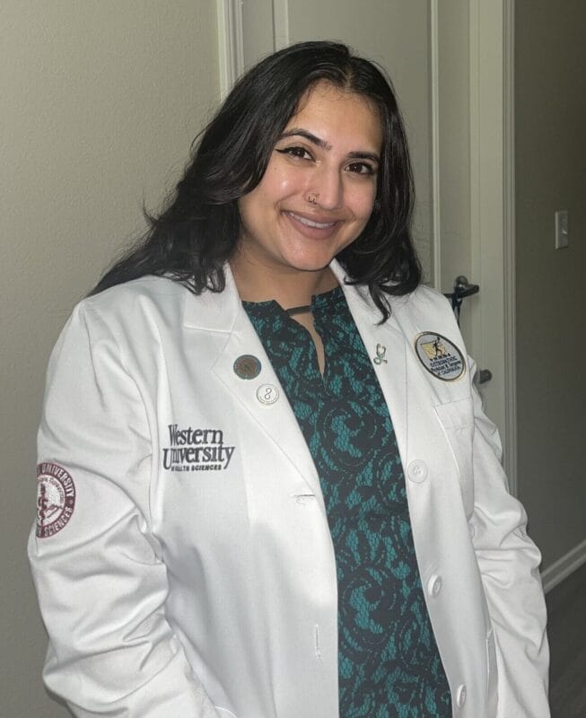 Woman wearing a white lab coat with "Western University" and other logos, standing indoors and smiling at the camera.