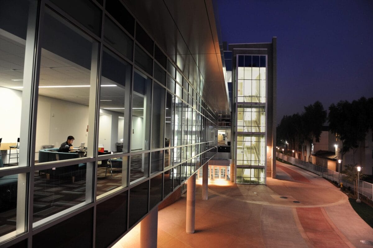 Modern building at night with large glass windows; a person works alone inside a brightly lit office while the exterior and surrounding area are dimly lit.