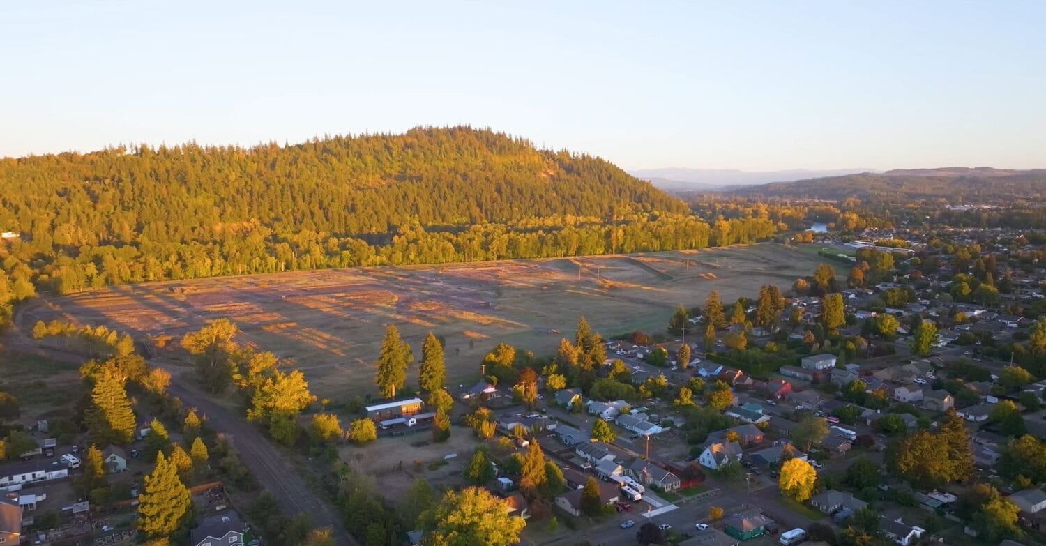 Aerial view of a large empty field bordered by trees and residential houses, with a forested hill in the background under a clear sky.